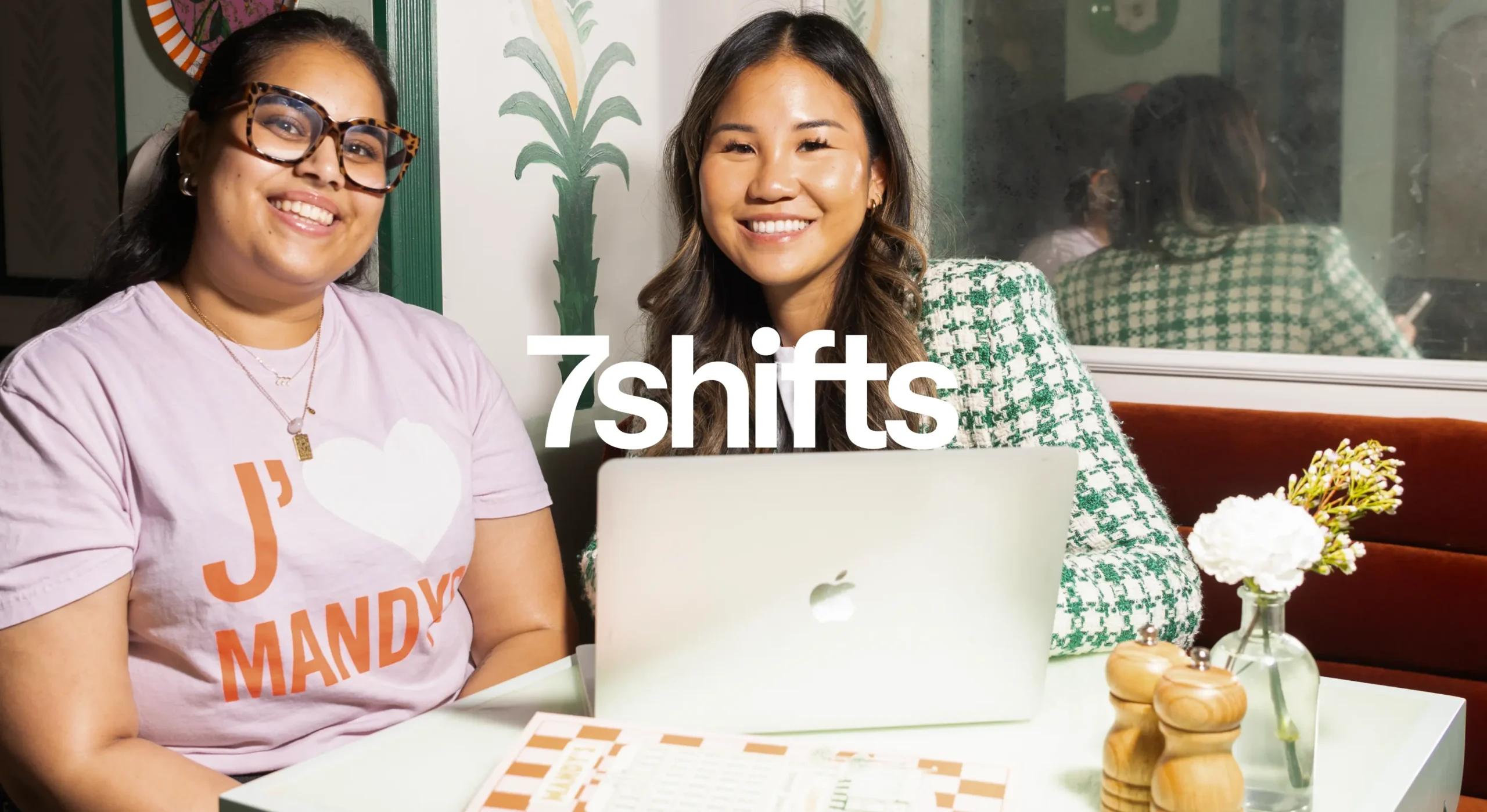 Image of two women smiling behind a laptop in a restaurant setting, white 7shifts logo on top