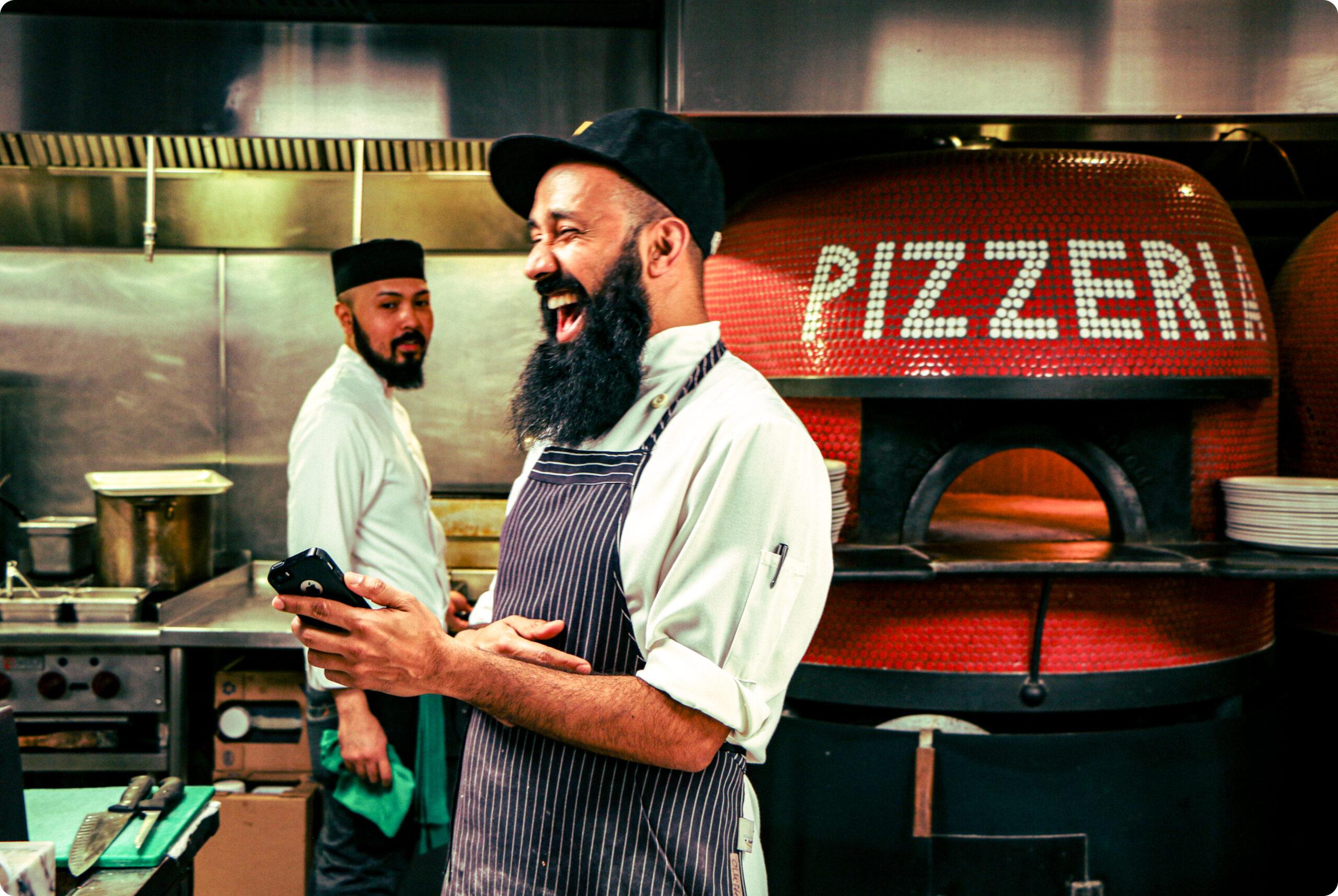 Bearded man laughing in pizzeria restaurant kitchen, holding a mobile phone